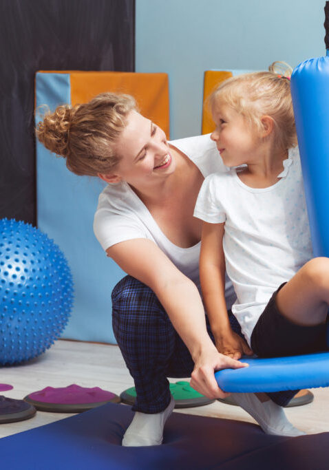 Young physiotherapist seesawing the girl on a blue swing